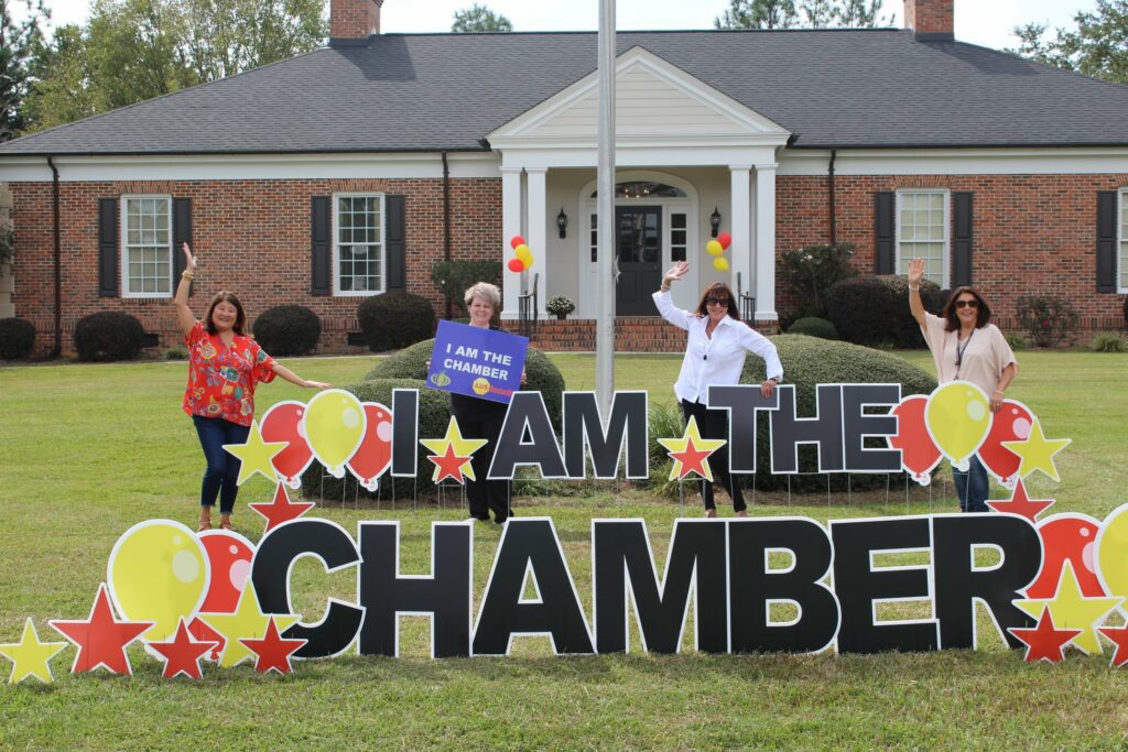 Image is of a large house with a sign that says, "I Am the Chamber," and people in front of it.