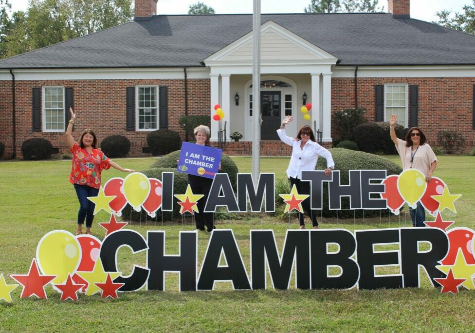 Image is of a large house with a sign that says, "I Am the Chamber," and people in front of it.